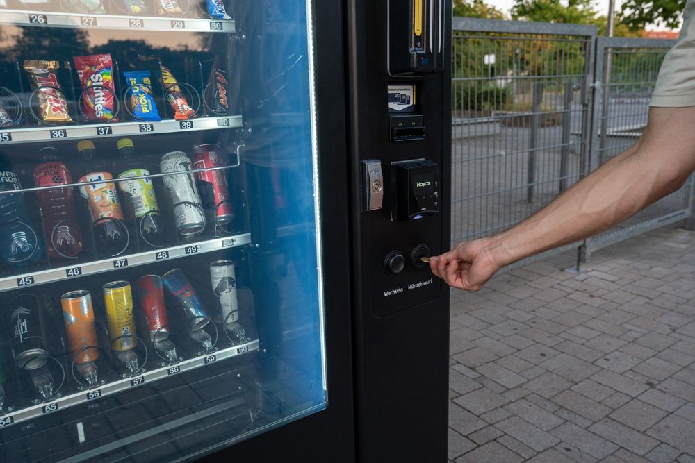 Hand am Münzrückgabeknopf eines Snackautomaten, Sortiment mit Preisschildern im Hintergrund