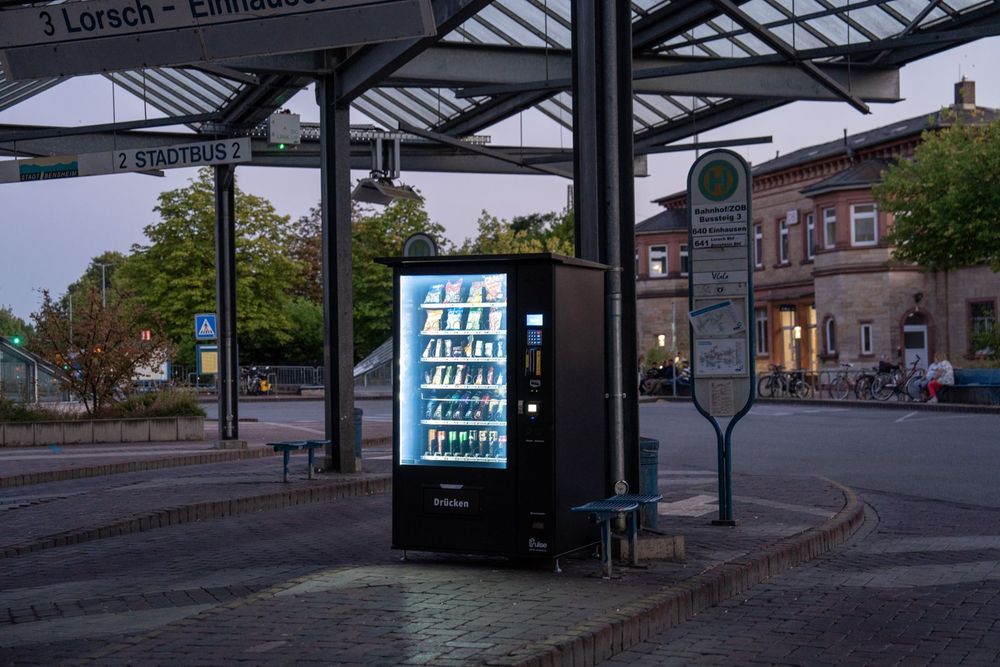 Pulse PV1100 Snackautomat am Busbahnhof in der Abenddämmerung, Bahnhofsgebäude im Hintergrund